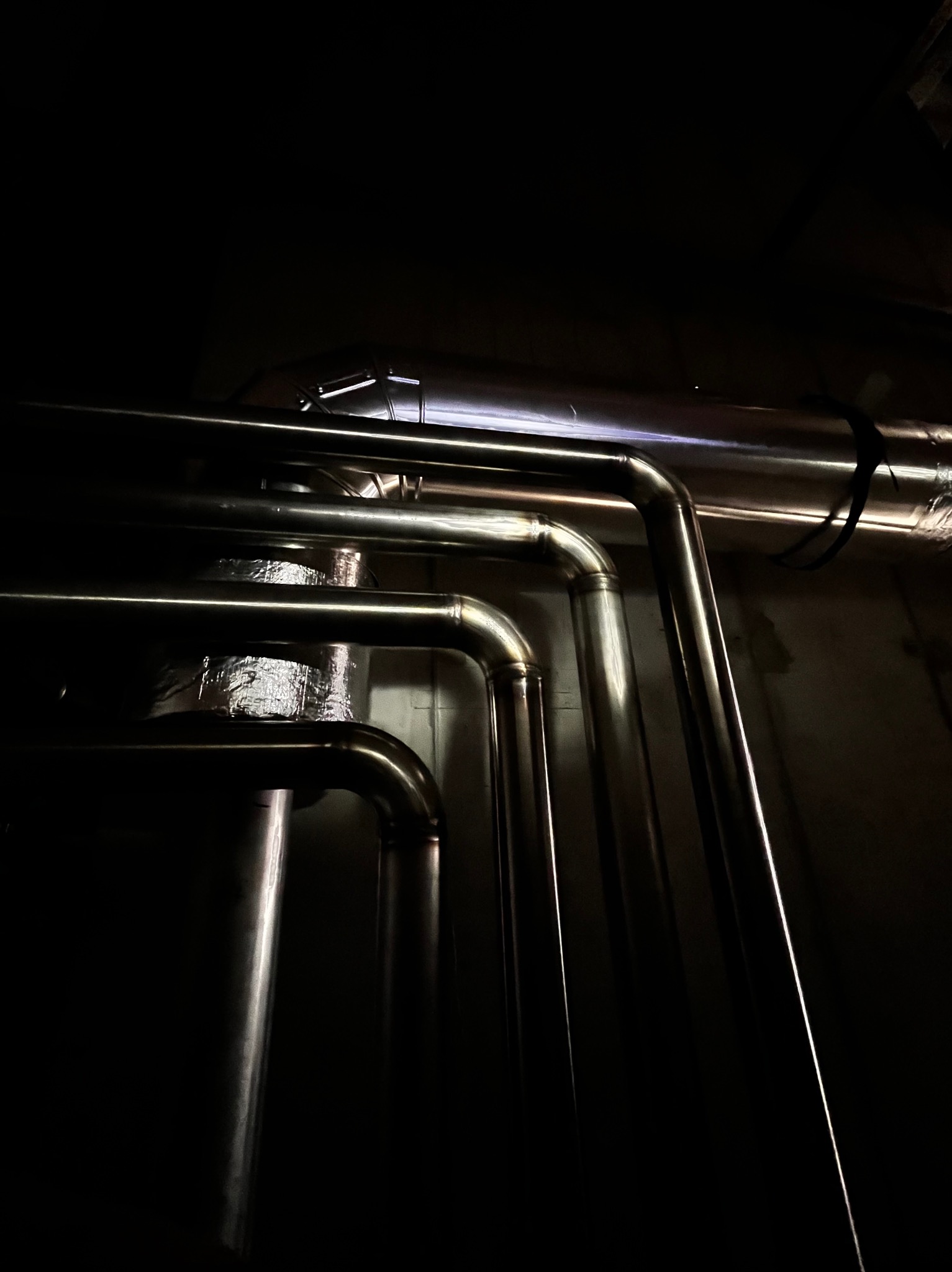 Instalacja z rur ze stali nierdzewnej, widoczne złącza kolankowe i izolacja termiczna owinięta folią aluminiową, w słabo oświetlonym pomieszczeniu technicznym.