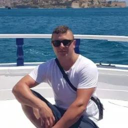 Mężczyzna w okularach przeciwsłonecznych siedzi na pokładzie łodzi, w tle widok na morze i starożytną fortecę na wyspie.