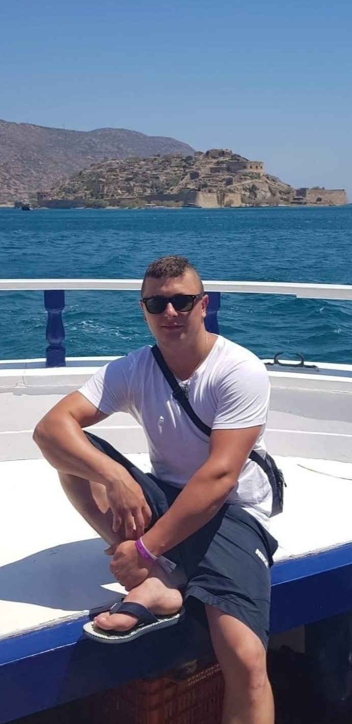 Mężczyzna w okularach przeciwsłonecznych siedzi na pokładzie łodzi, w tle widok na morze i starożytną fortecę na wyspie.