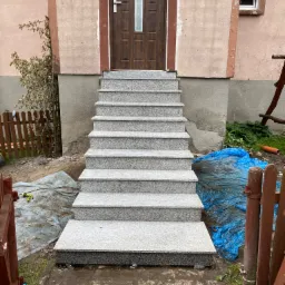 Granitowe schody zewnętrzne prowadzące do brązowych drzwi wejściowych, widoczne ślady prac budowlanych wokół.