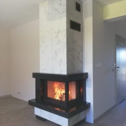 Kominki-Sulęcin Stefan Zarychta - Nowoczesny kominek narożny z obudową z białych płytek imitujących marmur, czarny wkład kominkowy z widocznym płomieniem, kratki wentylacyjne w obudowie.