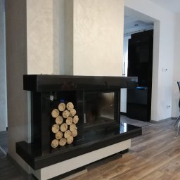 Kominki-Sulęcin Stefan Zarychta - Nowoczesny kominek z czarnego kamienia z ułożonym drewnem opałowym, w tle jasna ściana z dekoracyjną fakturą i fragment czarnej lodówki.