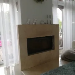 Nowoczesny kominek z obudową z jasnego kamienia, dekorowany figurkami i wiankiem, na tle białych zasłon i błyszczącej podłogi.