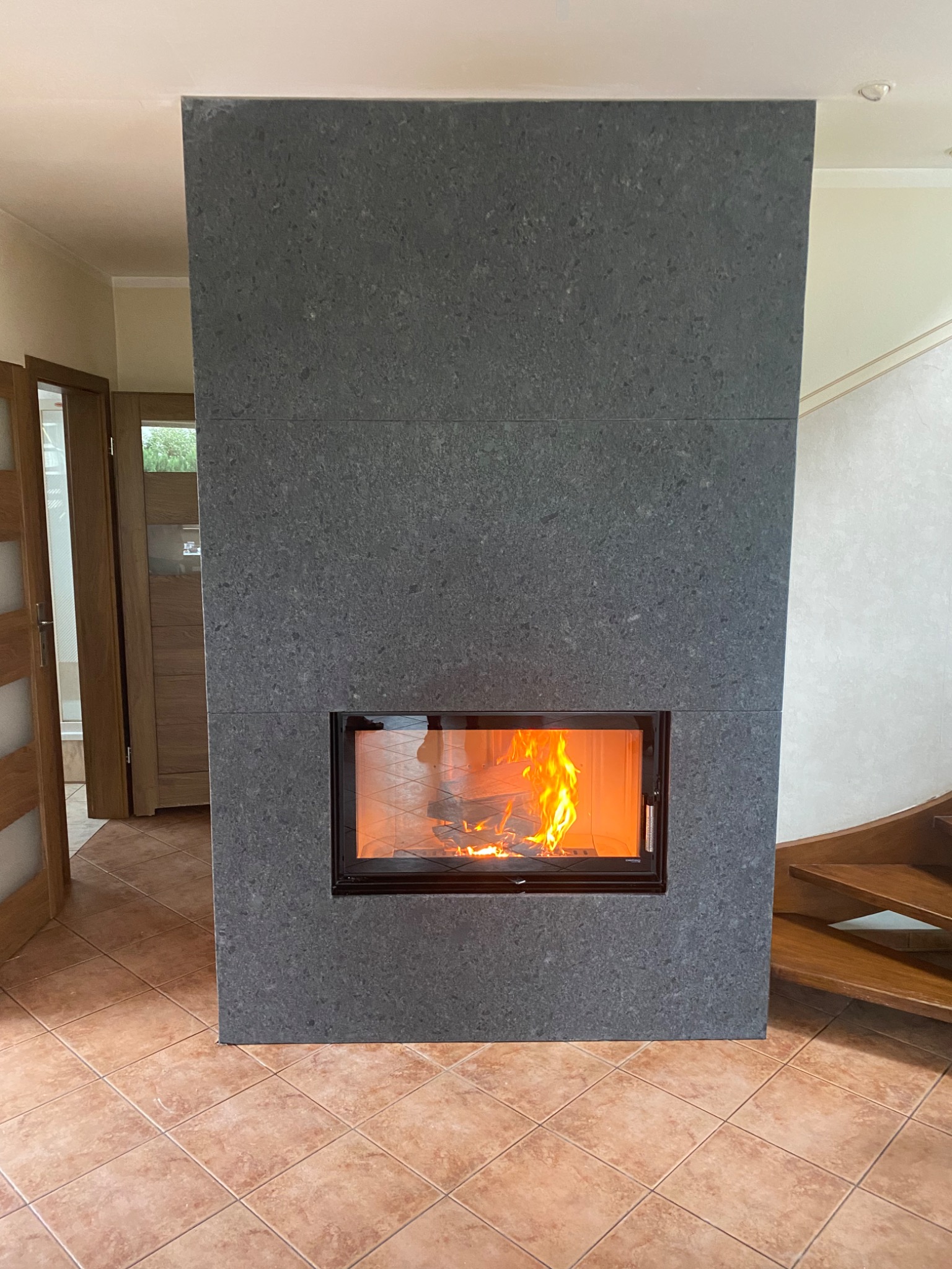Nowoczesny kominek z obudową z szarego kamienia, widoczne płomienie za szybą, w tle drewniane schody i drzwi.