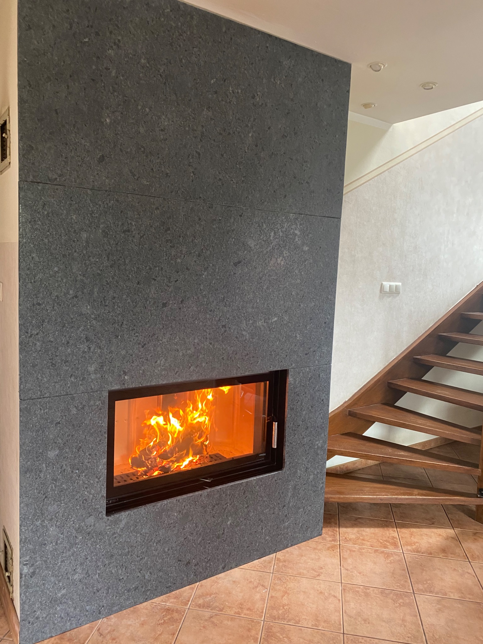 Nowoczesny kominek z obudową z szarego kamienia, płomienie tańczące za szybą, w tle drewniane schody i ceramiczna podłoga.