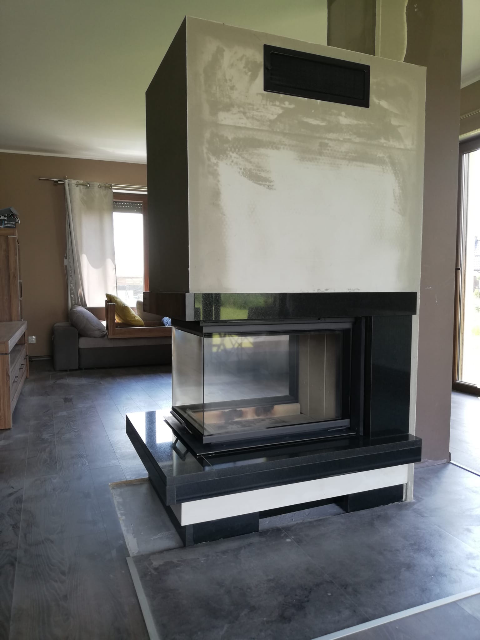 Nowoczesny kominek z czarnym obramowaniem i szklanym frontem w trakcie montażu, ściana surowa, widoczne niedociągnięcia wykończeniowe, podłoga w ciemnym odcieniu drewna.
