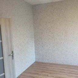 Pusty pokój po remoncie: ściany pokryte jasną tapetą strukturalną, drewnopodobna podłoga, widoczne drzwi z szarą ościeżnicą i srebrną klamką.