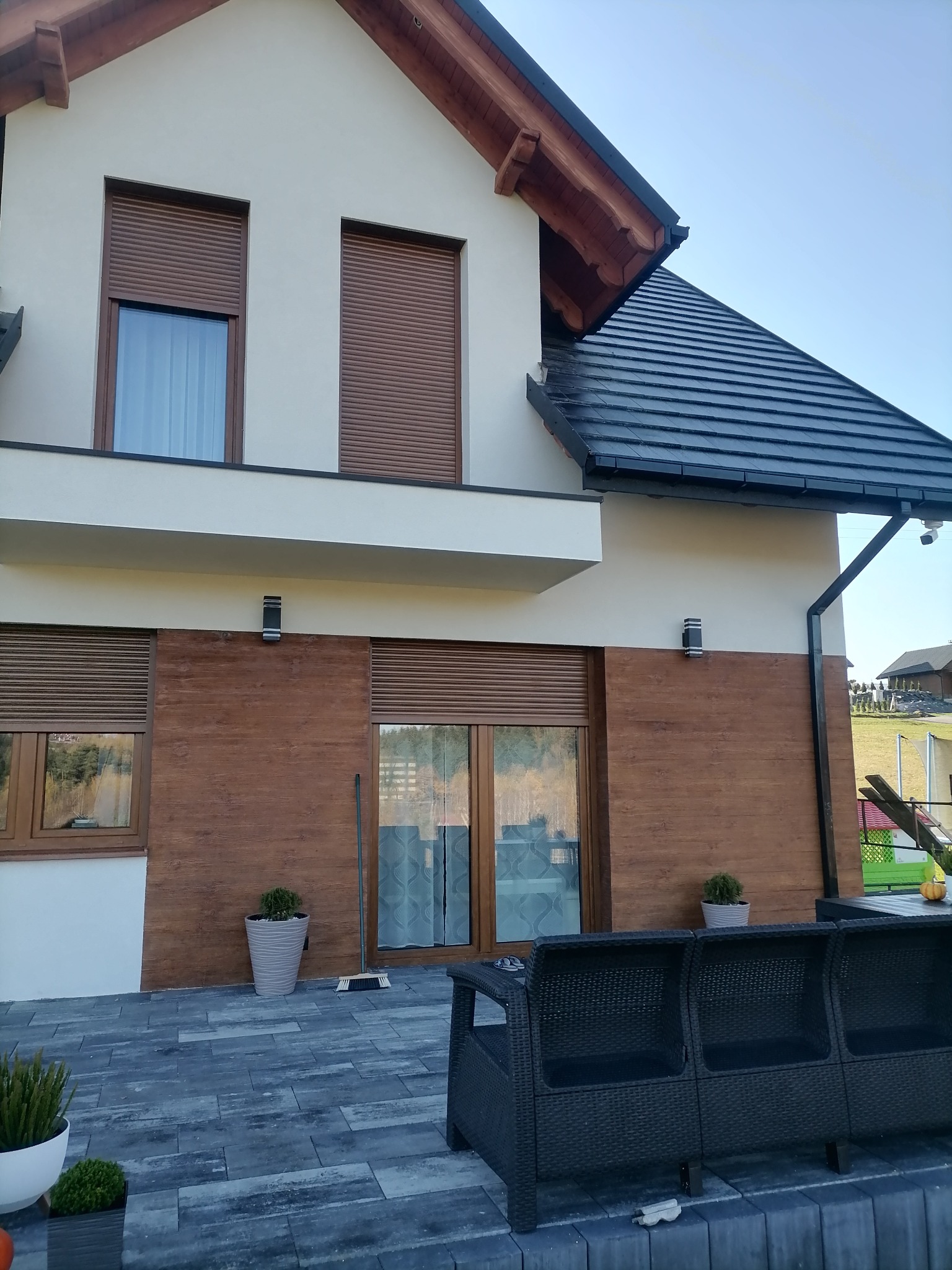 Elewacja domu z drewnopodobną okładziną, roletami zewnętrznymi i czarną dachówką, widok na taras z meblami ogrodowymi.