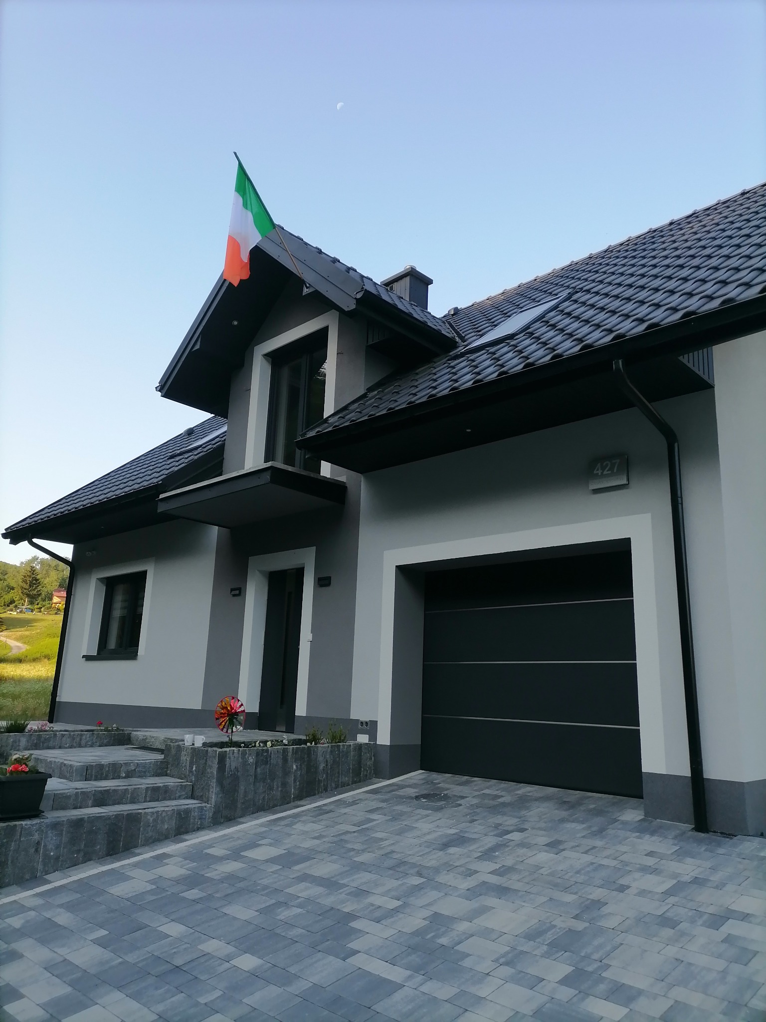 Nowoczesny dom jednorodzinny z szarą elewacją, ciemną dachówką i garażem, na dachu powiewa flaga Irlandii, widoczne kostka brukowa na podjeździe i schody wejściowe