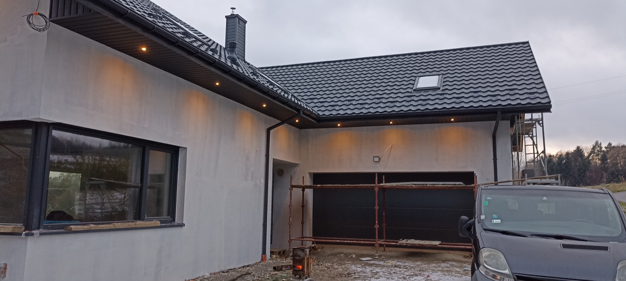 Dom jednorodzinny w trakcie prac elewacyjnych, z widocznymi rusztowaniami, świeżo nałożonym tynkiem, oświetleniem zewnętrznym pod dachem i nową stolarką okienną.