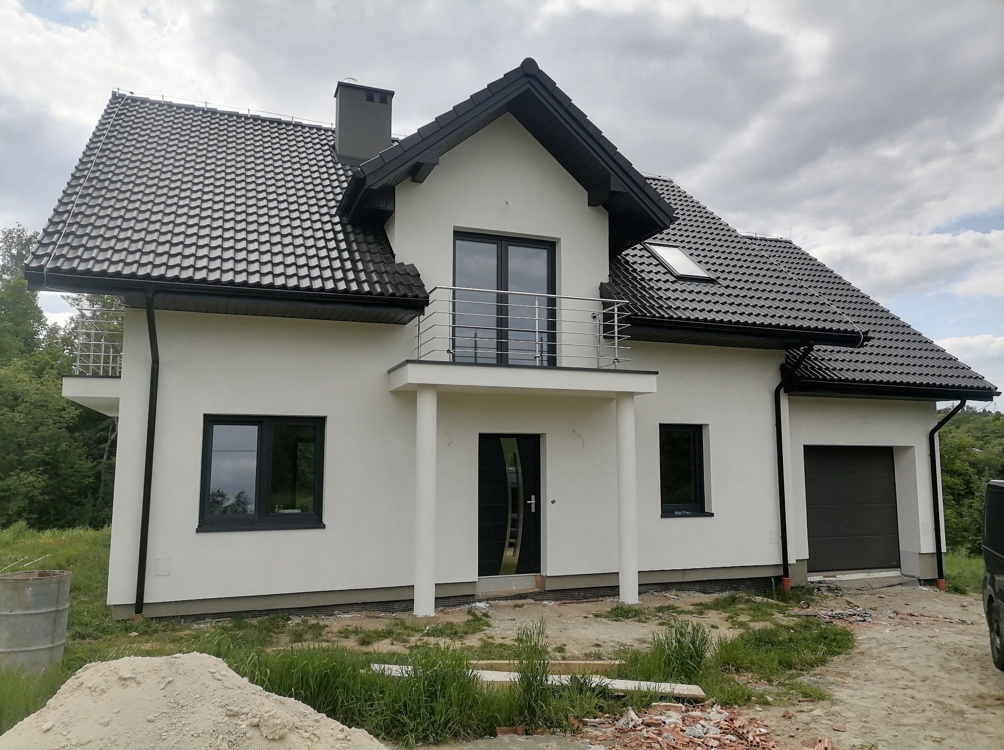 Nowoczesny dom jednorodzinny z białą elewacją, czarnym dachem z dachówki, balkonem i otwartymi drzwiami wejściowymi, otoczony gruzem i stertą piasku.