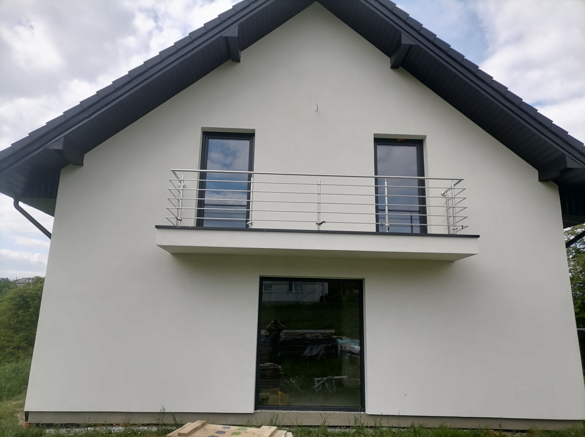 Wykończona elewacja domu jednorodzinnego z balkonem, widoczne okna z ciemnymi ramami, biała fasada, ciemna dachówka i stalowa balustrada balkonu.
