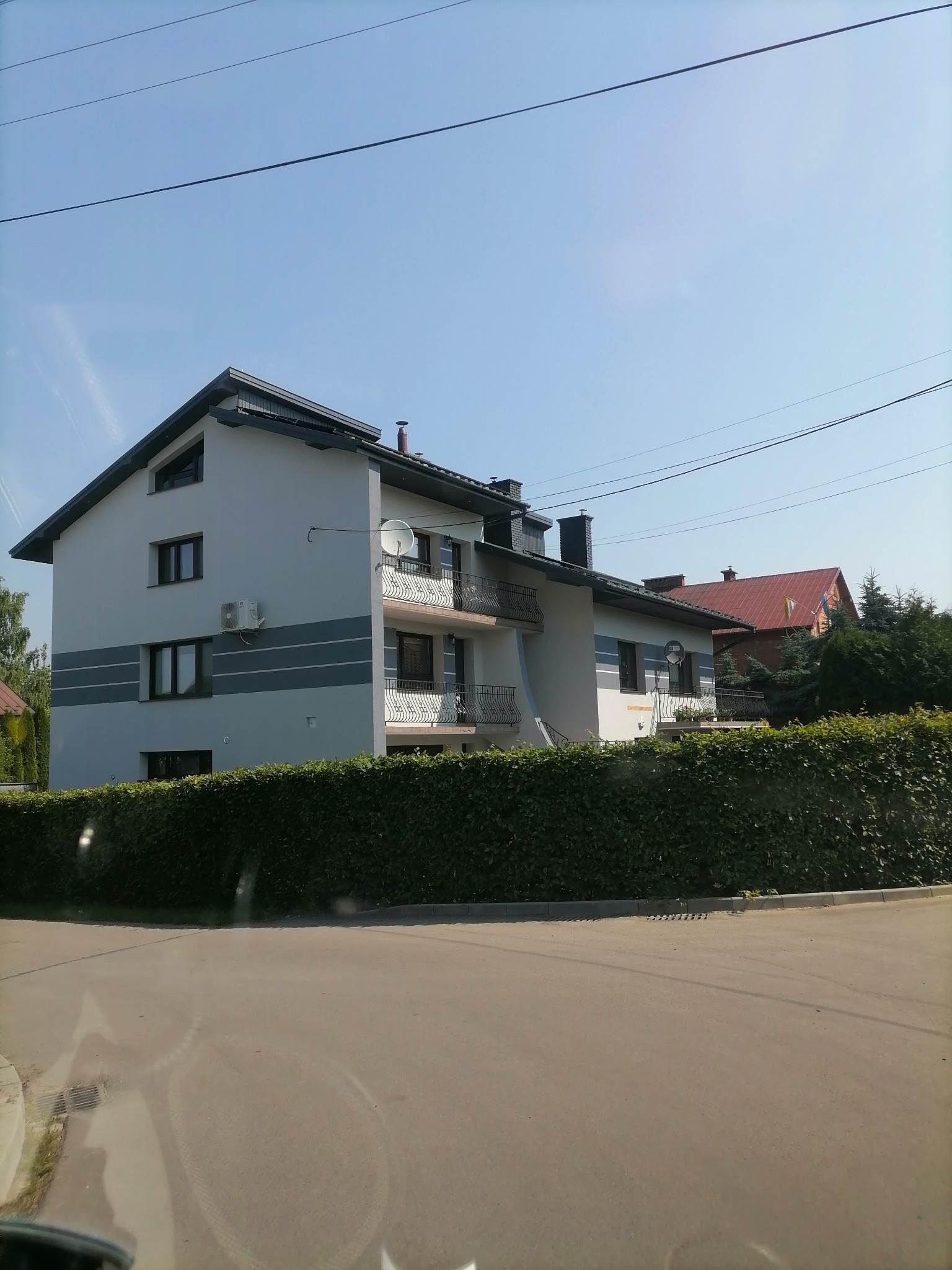 Dwupiętrowy dom z jasnoszarą elewacją, ozdobiony ciemnoszarymi pasami, z balkonami z kutymi balustradami i widoczną jednostką klimatyzacji na ścianie, sfotografowany z poziomu ulicy za żywopłotem.