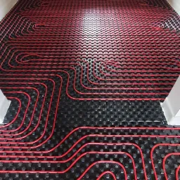 Czerwone rury ogrzewania podłogowego ułożone na czarnej macie w jasnym pomieszczeniu z oknem dachowym, widoczne stopy instalatora w dolnej części kadru.