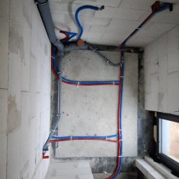 Usługi Hydrauliczne Sebastian Kutowski - Surowy stan instalacji wodnej i kanalizacyjnej w pomieszczeniu z bloczków, widoczne rury w kolorze szarym, niebieskim i czerwonym, przymocowane do ścian i sufitu.