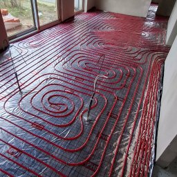 Usługi Hydrauliczne Sebastian Kutowski - Czerwone rury ogrzewania podłogowego ułożone spiralnie na folii izolacyjnej w surowym wnętrzu z oknami. Widoczne elementy montażowe i fragmenty ścian.