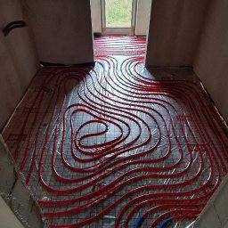 Usługi Hydrauliczne Sebastian Kutowski - Czerwone rury ogrzewania podłogowego ułożone na siatce w surowym wnętrzu z oknem. Widok z góry na instalację przed wylewką.