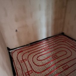 Usługi Hydrauliczne Sebastian Kutowski - Ułożona instalacja ogrzewania podłogowego w pomieszczeniu z surowymi ścianami. Czerwone rurki tworzą spiralny wzór na srebrnej folii izolacyjnej.