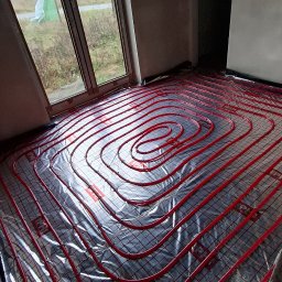 Usługi Hydrauliczne Sebastian Kutowski - Czerwone rury ogrzewania podłogowego ułożone spiralnie na folii izolacyjnej w jasnym pomieszczeniu z oknem. Widoczna siatka montażowa i fragment ściany.
