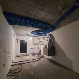 Wnętrze w trakcie instalacji: wiązka niebieskich rur pod sufitem, metalowe rury wentylacyjne i jednostka centralna przy ścianie, instalacje wodne i grzewcze poprowadzone w podłodze.
