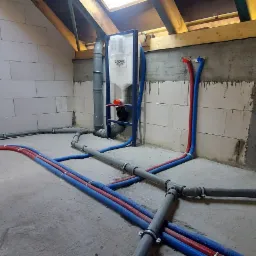 Instalacja wodno-kanalizacyjna w surowym stanie, widoczne rury w kolorze niebieskim i czerwonym prowadzone po podłodze i ścianach, częściowo zamontowany stelaż podtynkowy z białą spłuczką marki...