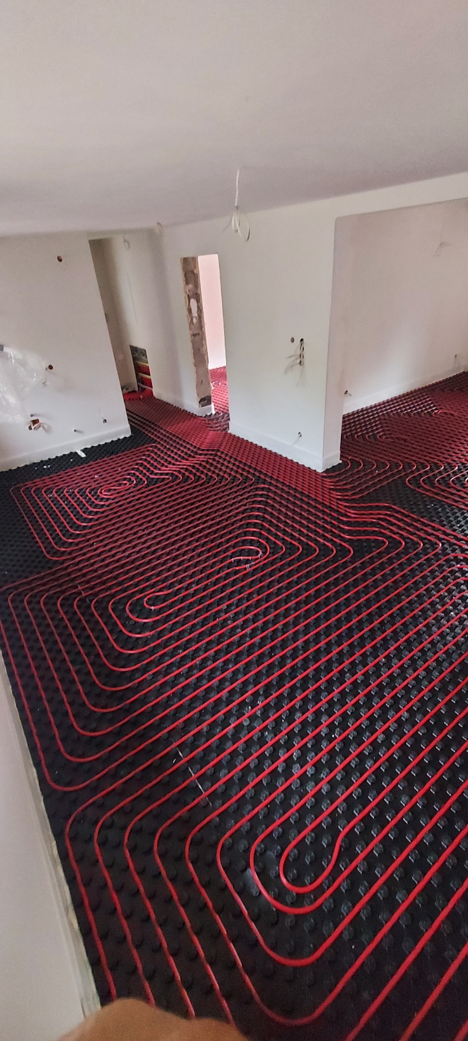 Czerwone rury ogrzewania podłogowego ułożone na czarnej macie izolacyjnej w surowym wnętrzu, widoczne przygotowane otwory na instalacje elektryczne w ścianach.