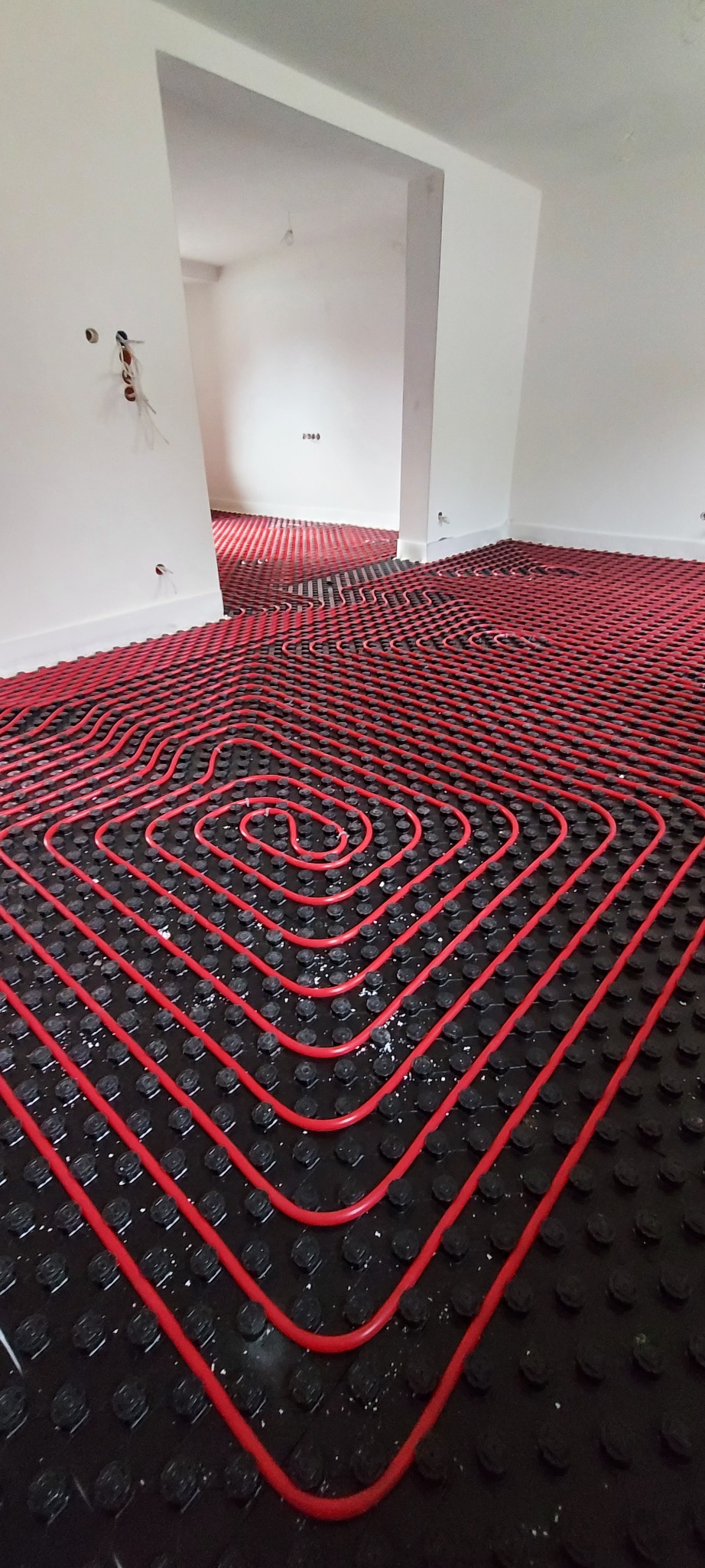 Czerwone rury ogrzewania podłogowego ułożone spiralnie na czarnej macie izolacyjnej w trakcie montażu w nowym, jasnym pomieszczeniu.