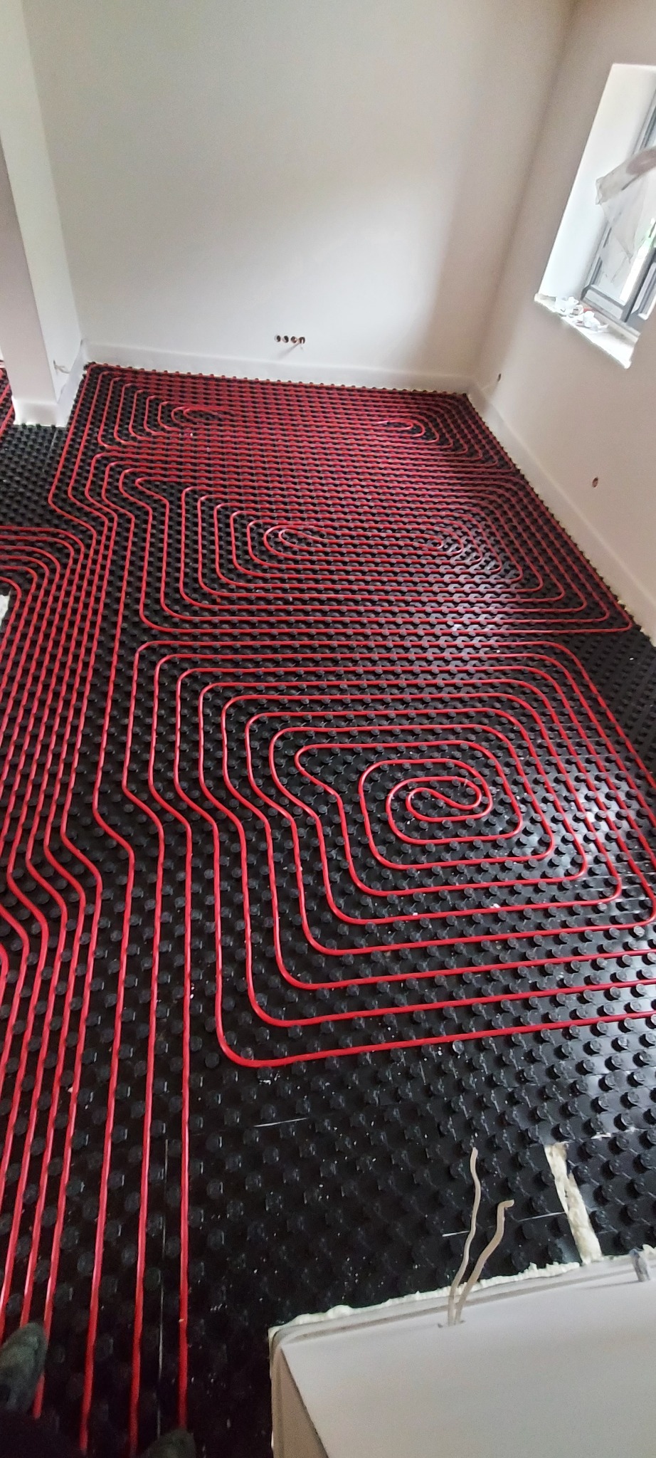 Czerwone rury ogrzewania podłogowego ułożone na czarnej macie z wypustkami w jasnym pomieszczeniu z oknem, widoczne fragmenty pianki montażowej przy ścianie.