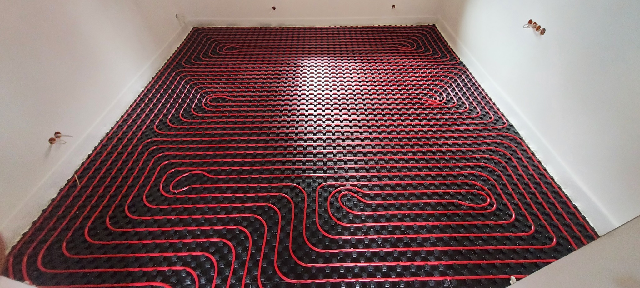Czerwone rury ogrzewania podłogowego ułożone na czarnej macie izolacyjnej w pustym pomieszczeniu z białymi ścianami, widoczne wyprowadzenia instalacji na ścianie.