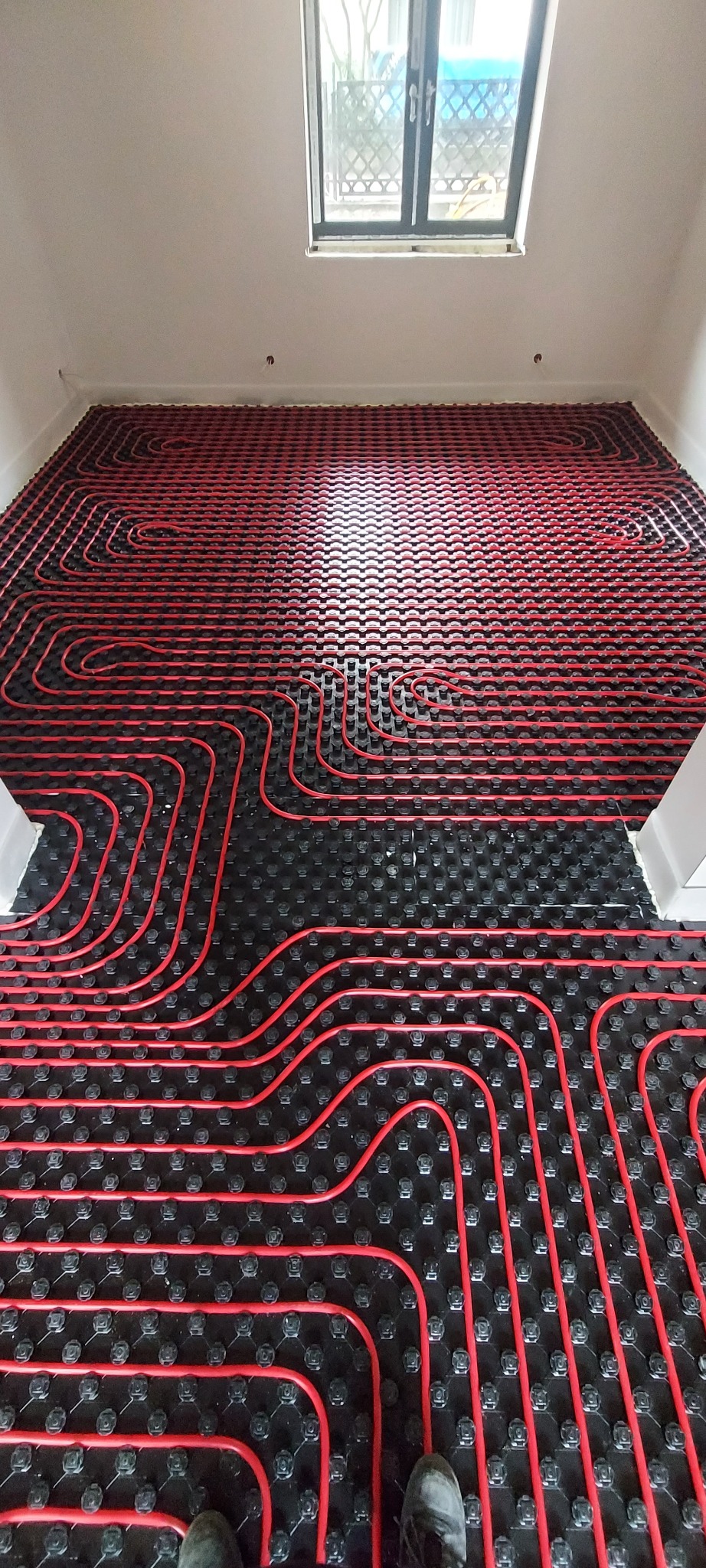 Czerwone rury ogrzewania podłogowego ułożone na czarnej macie w jasnym pomieszczeniu z oknem dachowym, widoczne stopy instalatora w dolnej części kadru.