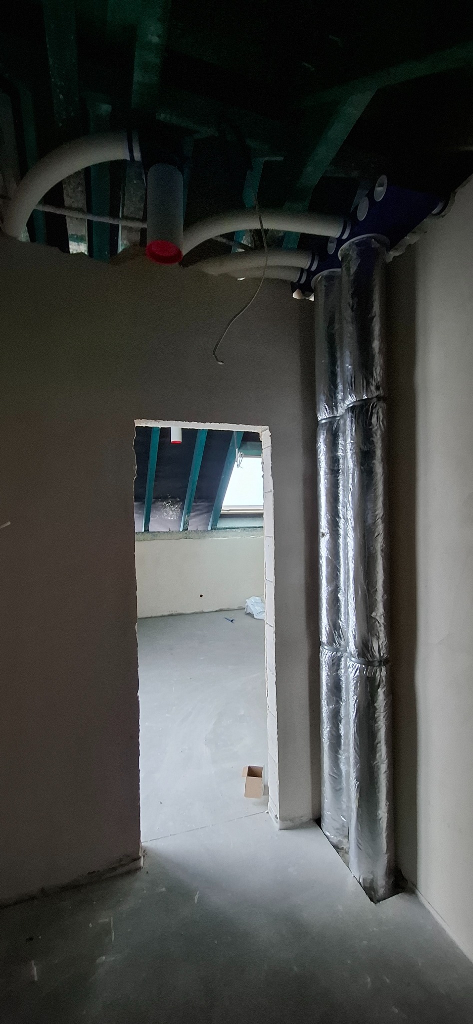 Dwa pionowe, izolowane przewody wentylacyjne w surowym wnętrzu. Widoczne białe rury pod sufitem i otwór drzwiowy z widokiem na poddasze. Surowy stan wykończenia.