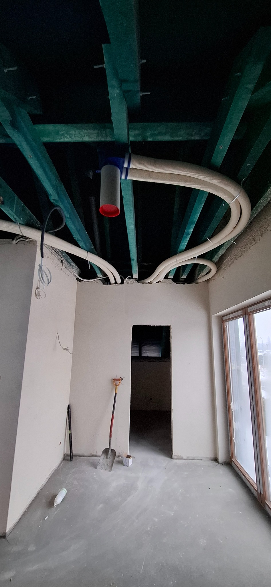 Instalacja wentylacyjna z rurami karbowanymi pod sufitem z belek w surowym wnętrzu. Widoczna łopata i poziomica, okno z widokiem na miasto.