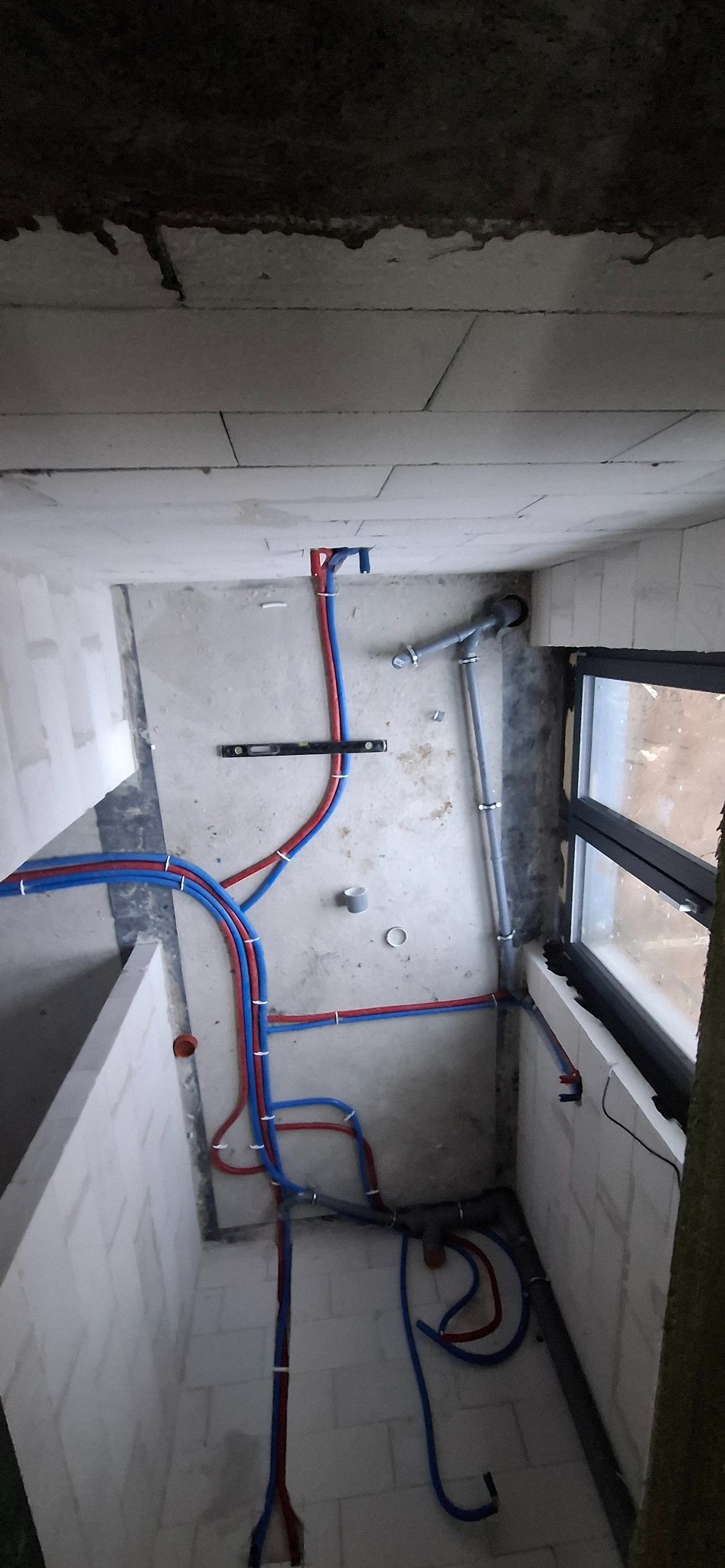 Widok z góry na fragment wnętrza w trakcie prac instalacyjnych: rury wodne (czerwone i niebieskie) oraz kanalizacyjne, poziomica na podłodze, okno w tle.