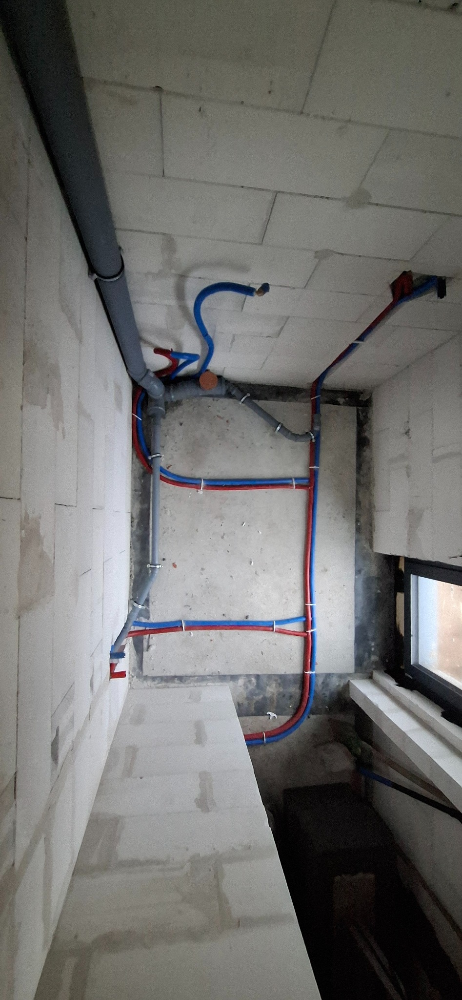 Surowy stan instalacji wodnej i kanalizacyjnej w pomieszczeniu z bloczków, widoczne rury w kolorze szarym, niebieskim i czerwonym, przymocowane do ścian i sufitu.
