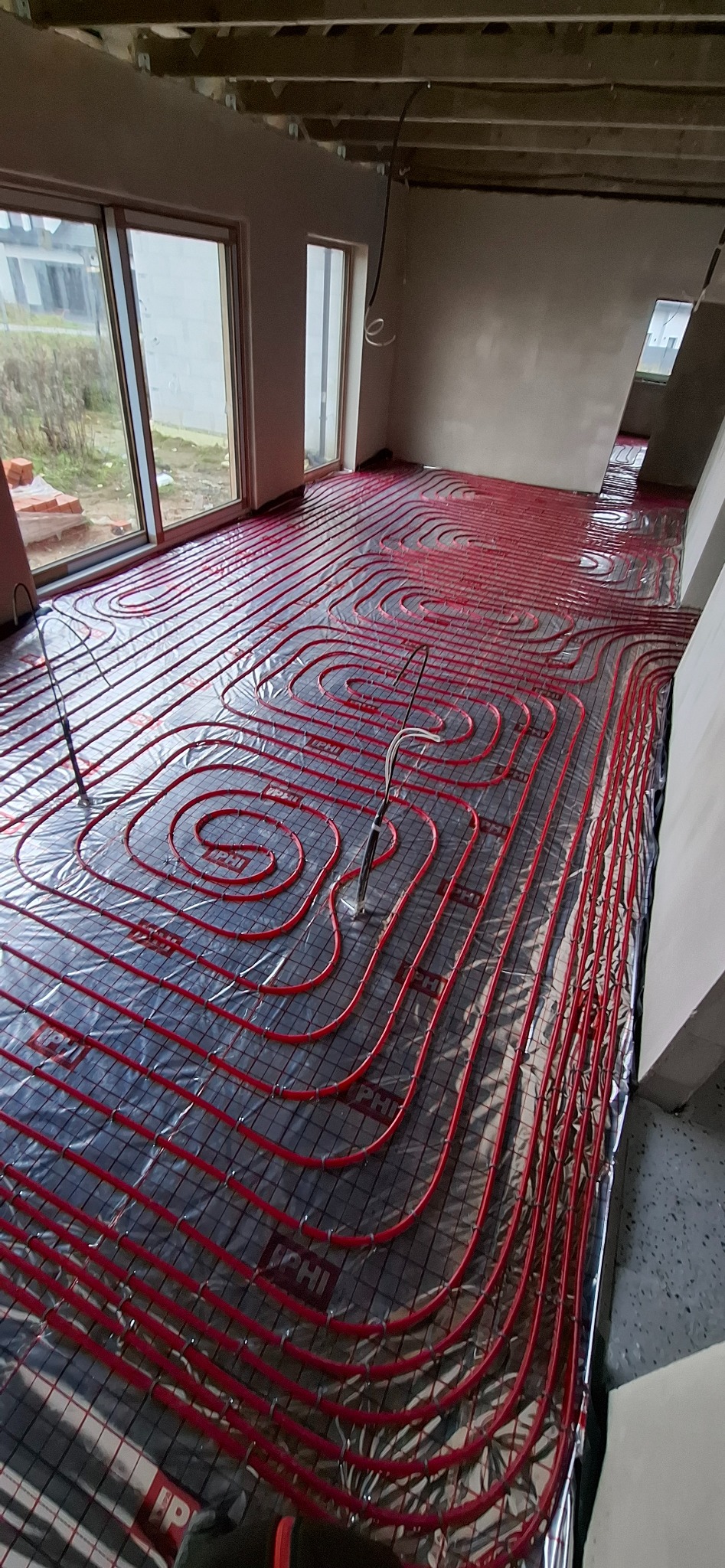 Czerwone rury ogrzewania podłogowego ułożone spiralnie na folii izolacyjnej w surowym wnętrzu z oknami. Widoczne elementy montażowe i fragmenty ścian.