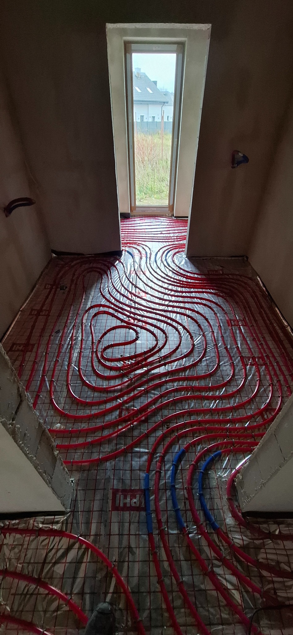 Czerwone rury ogrzewania podłogowego ułożone na siatce w surowym wnętrzu z oknem. Widok z góry na instalację przed wylewką.