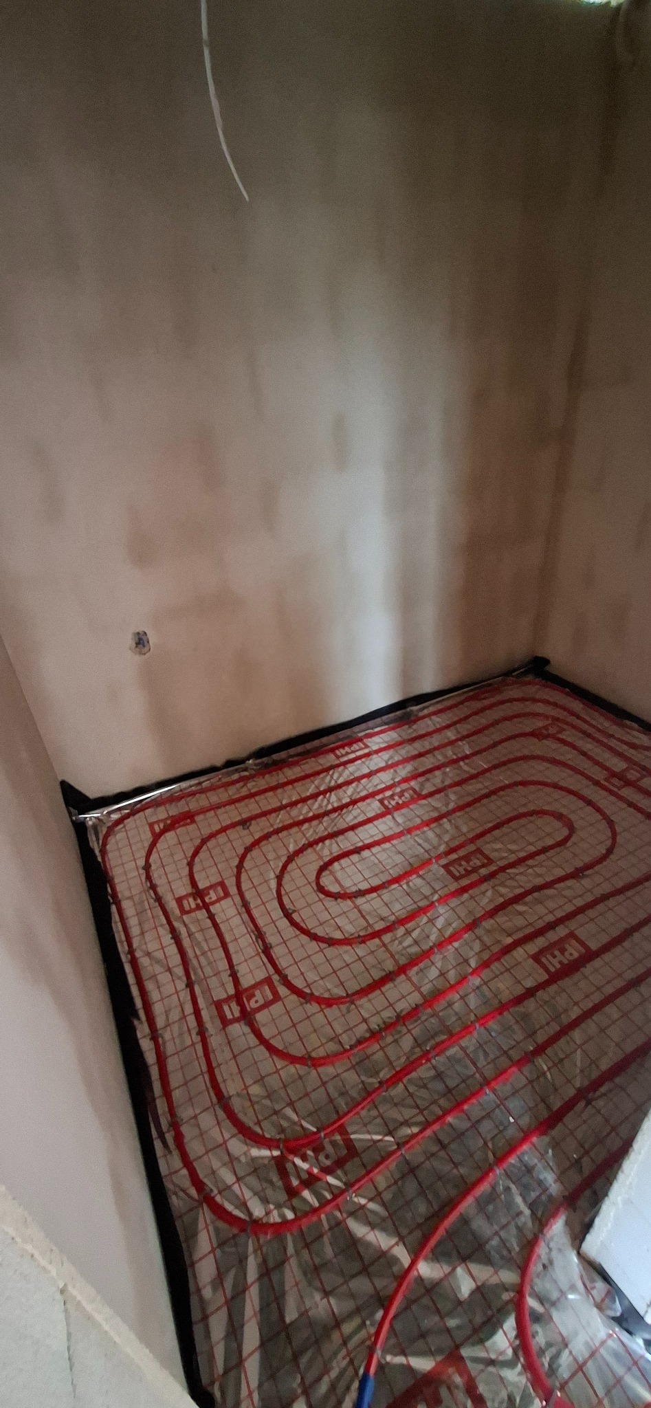 Ułożona instalacja ogrzewania podłogowego w pomieszczeniu z surowymi ścianami. Czerwone rurki tworzą spiralny wzór na srebrnej folii izolacyjnej.