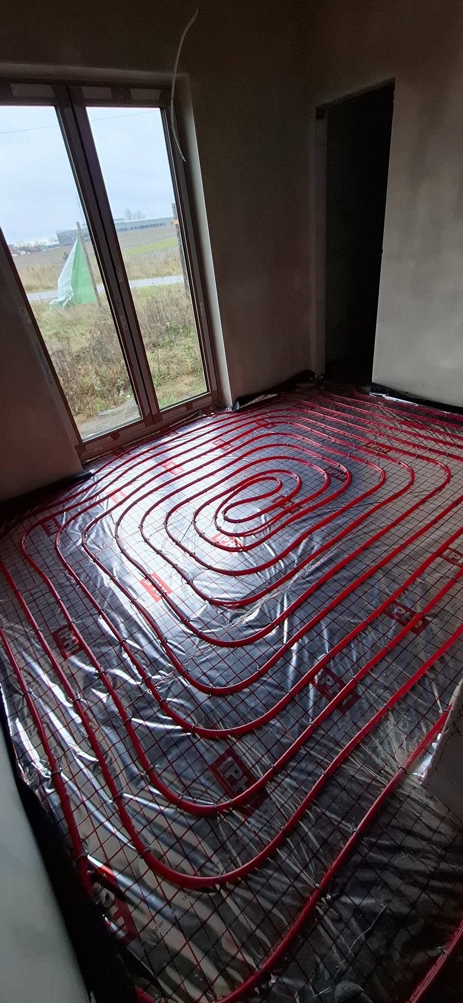 Czerwone rury ogrzewania podłogowego ułożone spiralnie na folii izolacyjnej w jasnym pomieszczeniu z oknem. Widoczna siatka montażowa i fragment ściany.