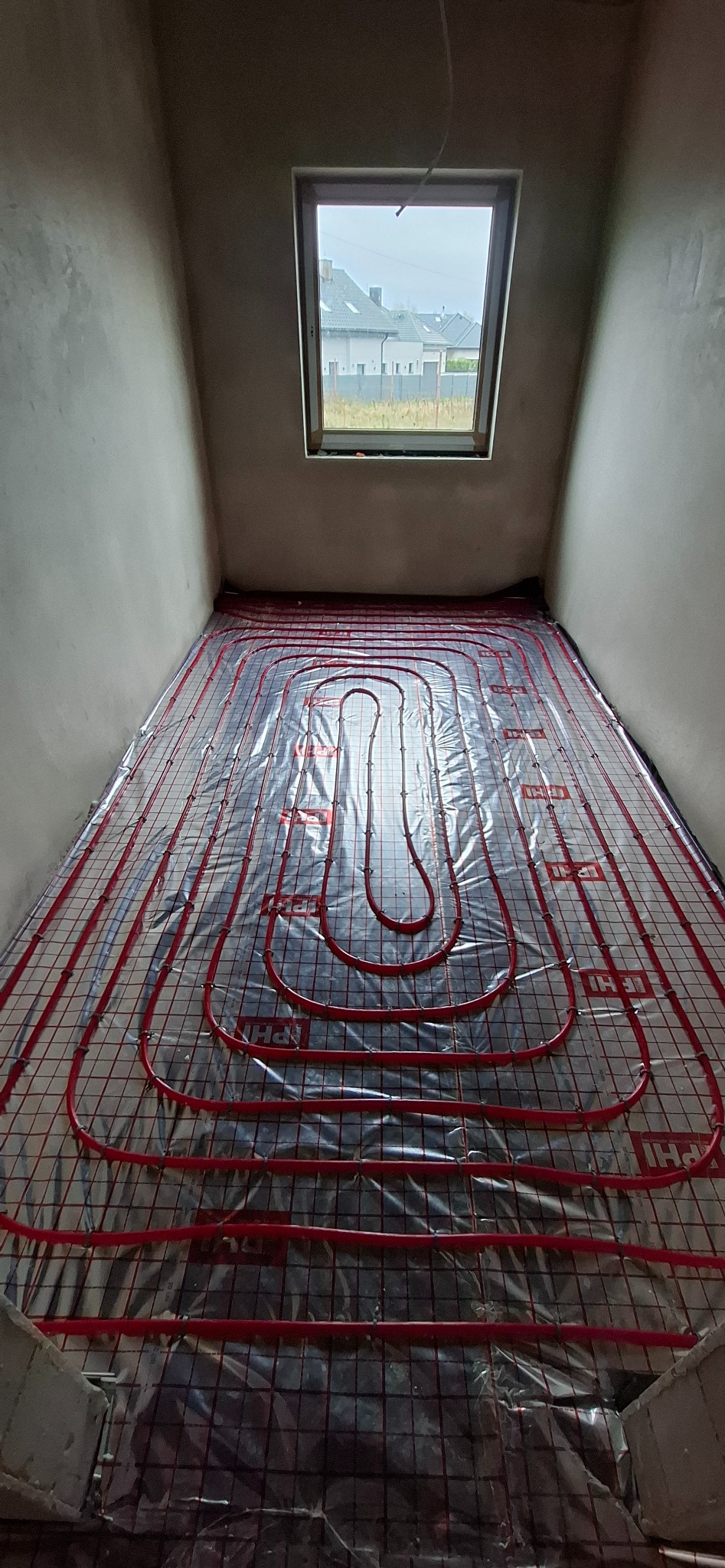 Czerwone rury ogrzewania podłogowego ułożone na folii izolacyjnej w surowym pomieszczeniu z oknem dachowym. Widok z góry na instalację przed wylaniem posadzki.