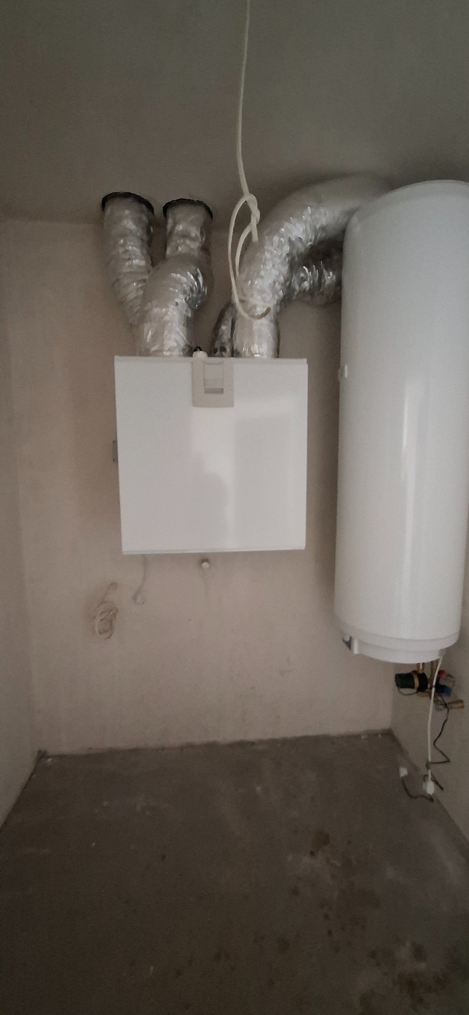 Biały bojler i rekuperator z aluminiowymi przewodami wentylacyjnymi na tle surowej ściany. Widoczne podłączenia elektryczne i hydrauliczne.