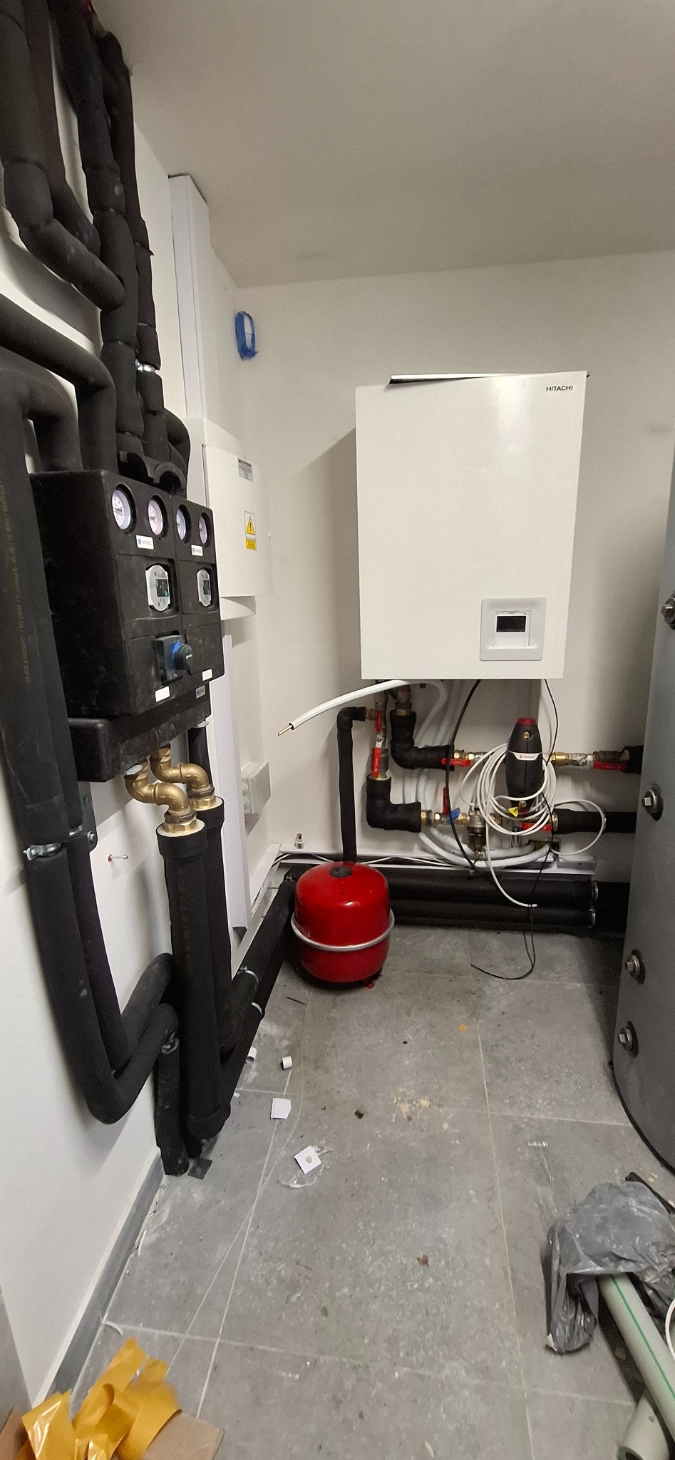 Biały piec CO marki Hitachi z czarnymi rurami w izolacji, czerwony zbiornik wyrównawczy i szary zasobnik. Widoczne manometry i instalacja elektryczna.