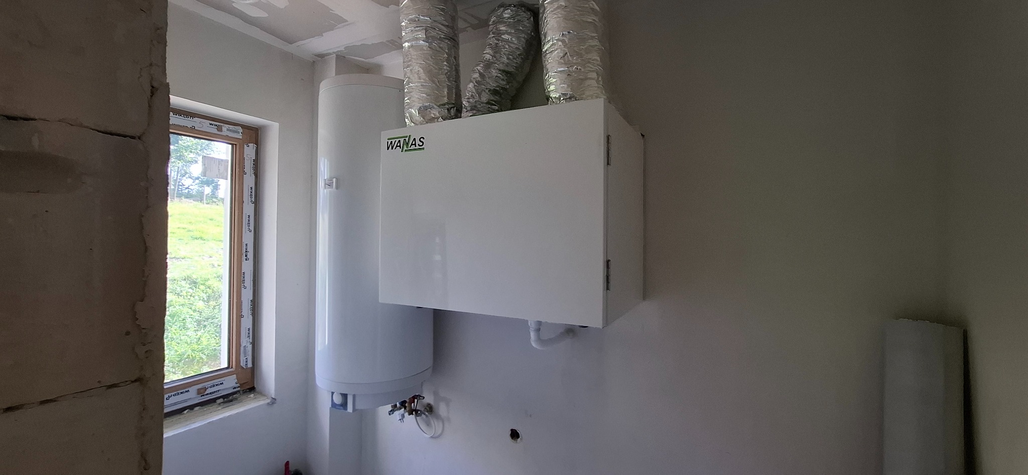 Biały bojler i rekuperator marki WANAS z aluminiowymi przewodami wentylacyjnymi w surowym wnętrzu, widoczne przyłącza hydrauliczne i okno z widokiem na zieleń.
