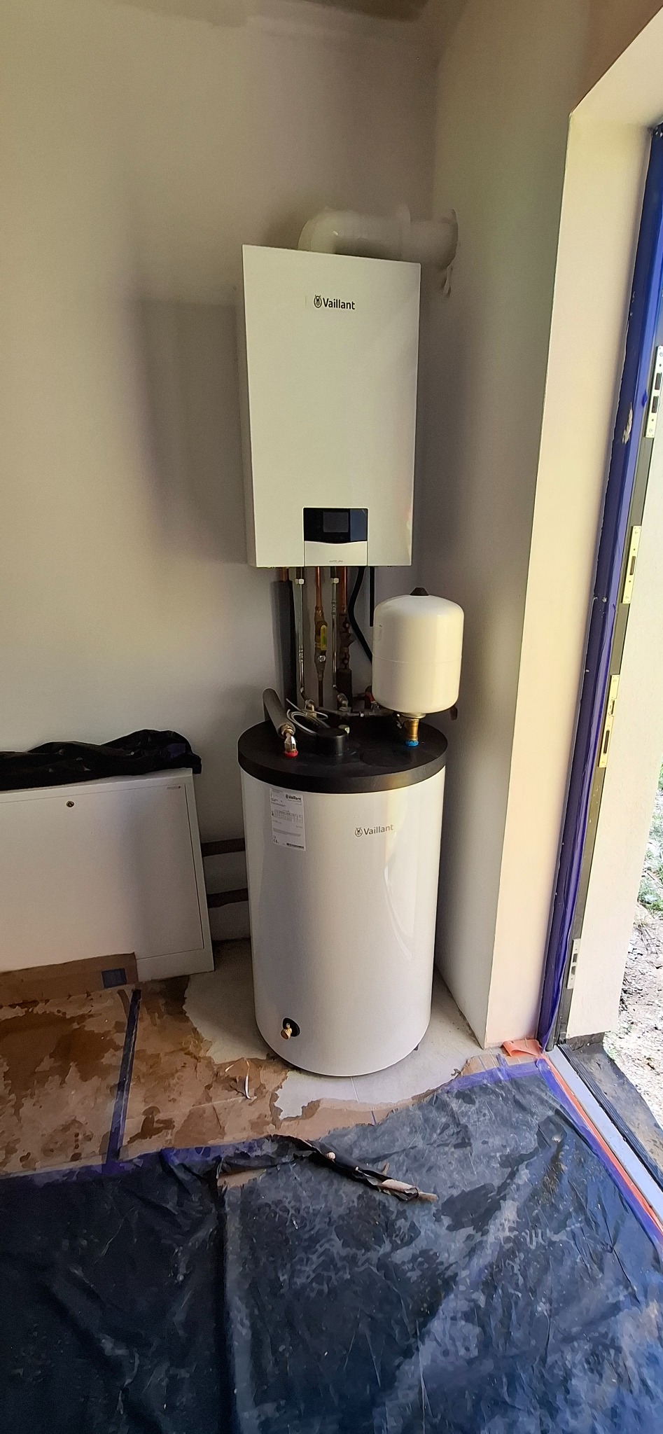 Biały, kondensacyjny piec gazowy Vaillant z zasobnikiem C.W.U. zamontowany w pomieszczeniu. Widoczne rury i elementy instalacji. Podłoga zabezpieczona folią.