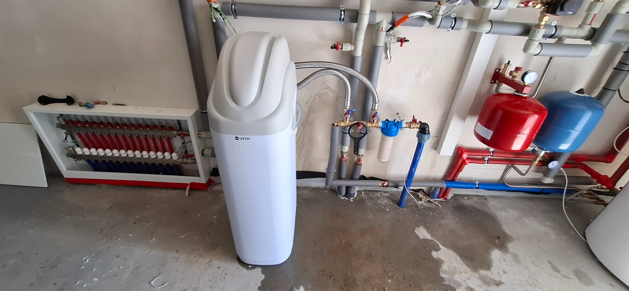 Biały zmiękczacz wody USTM na tle instalacji CO z rozdzielaczem podłogówki, zbiornikami wyrównawczymi i filtrem narurowym. Szare rury i betonowa posadzka.