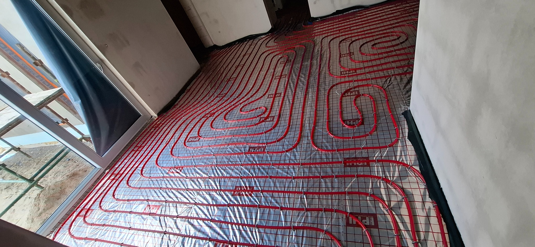 Czerwone rury ogrzewania podłogowego ułożone na folii, widoczne przez otwarte okno. Instalacja w trakcie realizacji, ściany surowe, brak wylewki.