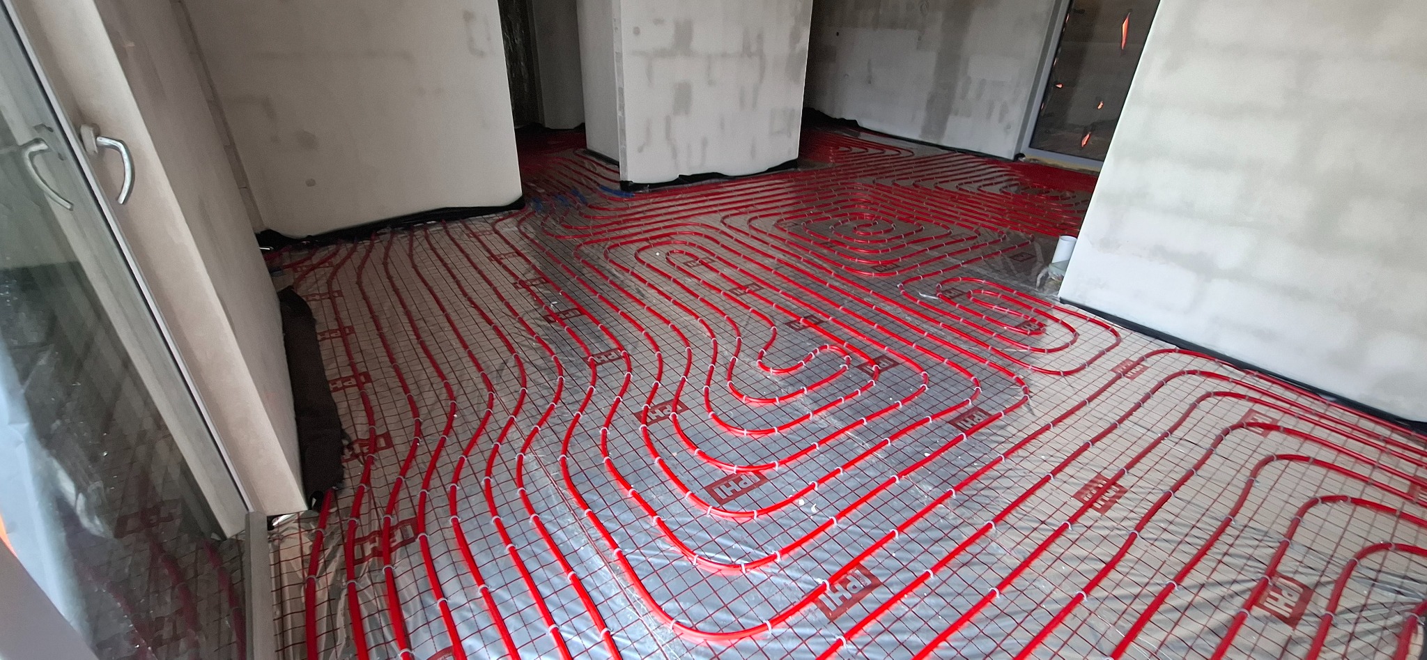 Czerwone rury ogrzewania podłogowego ułożone na srebrnej folii w surowym wnętrzu. Widoczne fragmenty ścian i okna. Charakterystyczny wzór ułożenia rur.