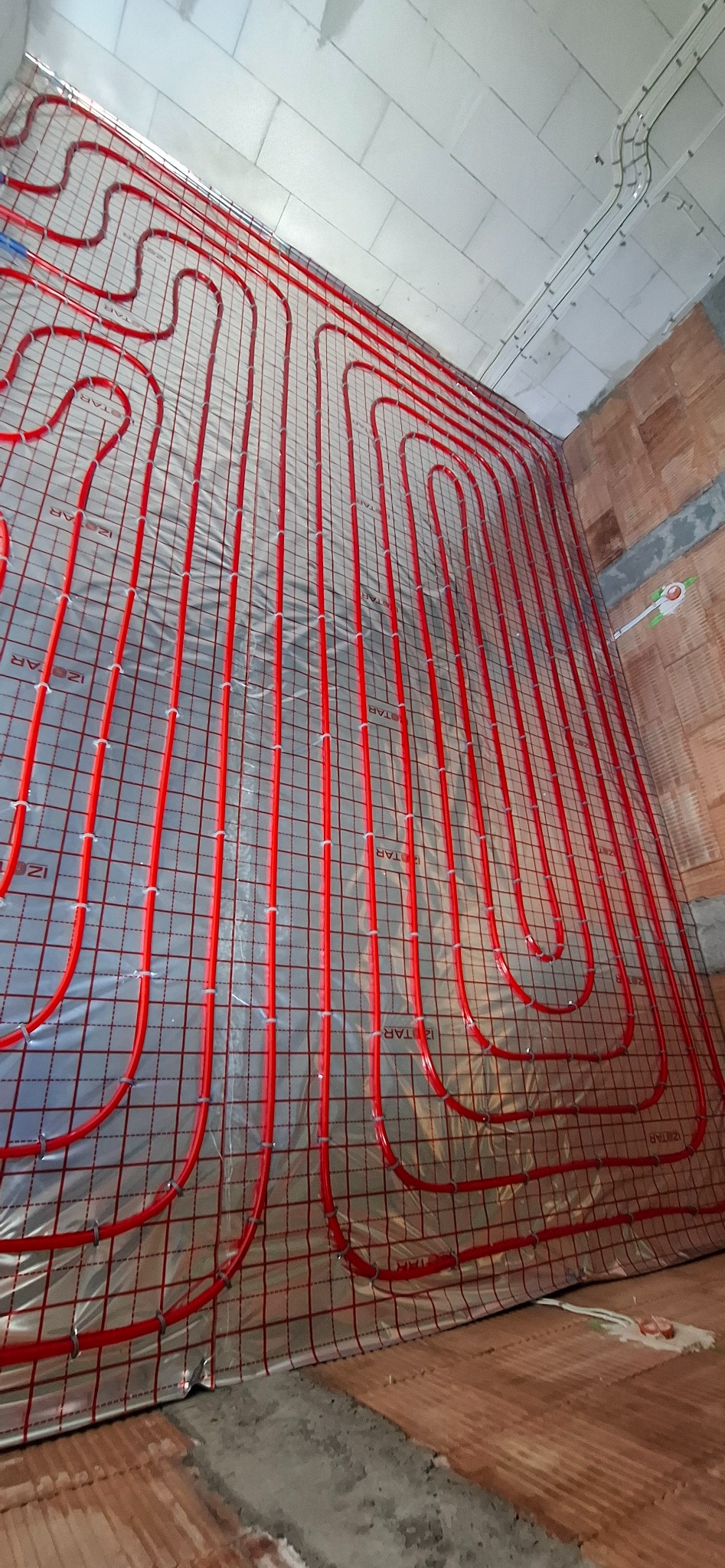 Czerwone rury ogrzewania podłogowego ułożone na folii z siatką, widok z góry, surowy stan wykończenia wnętrza, instalacja przed zalaniem posadzką.