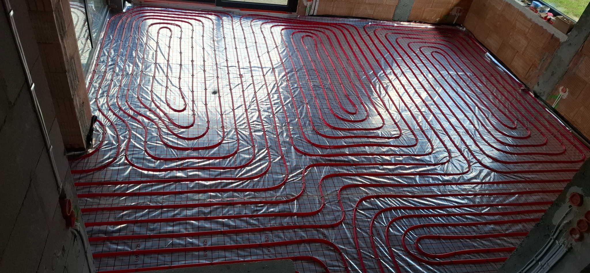 Czerwone rury ogrzewania podłogowego ułożone na srebrnej folii izolacyjnej z siatką, widok z góry, surowy stan budynku, przygotowanie do wylewki.