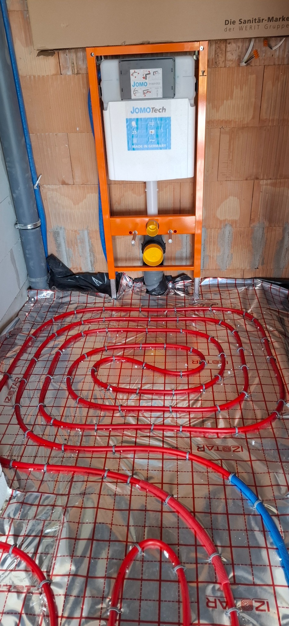 Instalacja ogrzewania podłogowego z czerwonymi rurami na folii izolacyjnej, widoczny stelaż podtynkowy WC JOMOtech i fragment instalacji wod-kan w surowym wnętrzu.