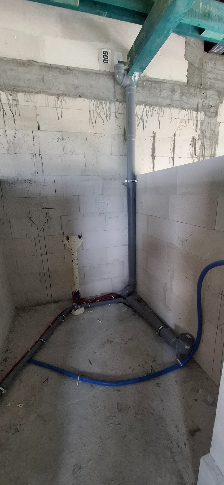 Surowy stan: pion kanalizacyjny z szarego PCV, rury wodne czerwone i niebieskie, pianka montażowa, ściany z betonu komórkowego. Instalacja w trakcie realizacji.